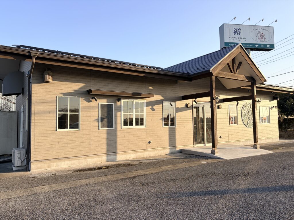 青空の下に立つ、平屋建ての独立型貸店舗の外観。木目調の外壁に三角屋根のエントランスが特徴的で、店舗前には広いアスファルトの駐車スペースが完備されている。