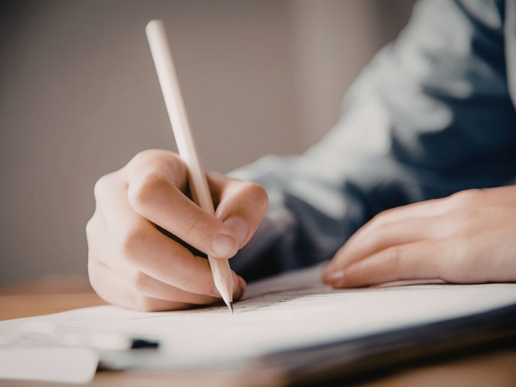 木製の机の上で、白い紙に鉛筆で文字や注意点を書き留めている人の手のクローズアップ。手元にピントが合っており、背景は柔らかくぼやけている。落ち着いたトーンのビジネスや学習、契約のイメージ写真。
