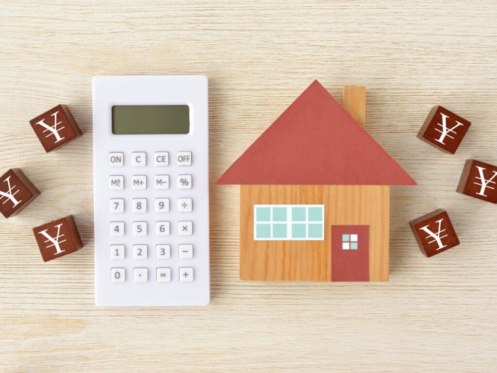 木製の家と電卓、周りに配置された円（¥）マークのブロック。不動産の相続税や評価額の計算イメージ。