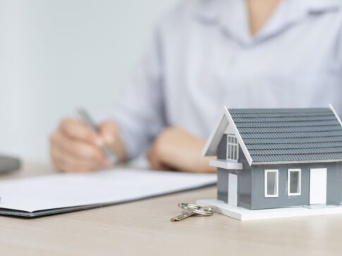 手前に一戸建ての模型と鍵、奥で書類にペンで記入する人の手元。市原市で相続した実家の売却・買取手続きのイメージ。