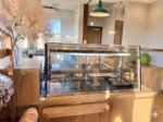 カフェや惣菜店の居抜き物件のカウンター周辺内装。中央に設置された2段式のガラス製冷蔵ショーケースと、その奥に見えるステンレス製の厨房シンクや作業スペース。木目調のレジカウンター、緑のペンダントライト、スモークツリーの装飾が調和した温かみのある店内。