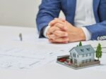 市原市での不動産の相続放棄をテーマに、机の上の住宅模型と図面、奥で相談に乗るスーツ姿の専門家の手元を写した写真