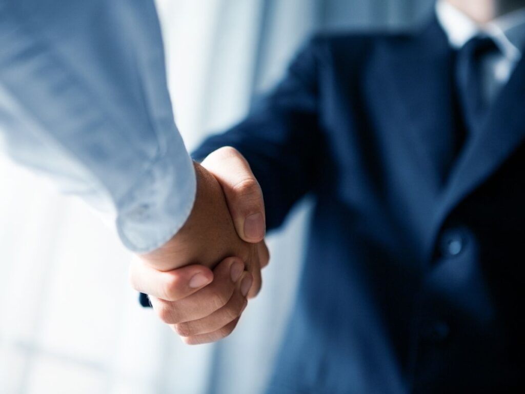 オフィスの会議室で、スーツを着た二人のビジネスパーソンが契約成立の握手を交わしている様子。