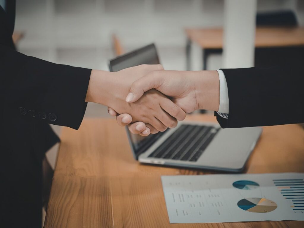 オフィスの木製デスクで、黒いスーツを着た2人のビジネスパーソンがしっかりと握手を交わしているクローズアップ写真。デスクの上には、開いたラップトップと、円グラフや棒グラフが描かれたビジネス文書が置かれている。ビジネスの合意やパートナーシップ成立の瞬間を捉えた画像。