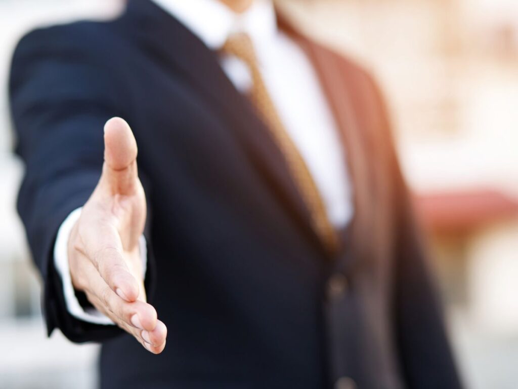 黒いビジネススーツとゴールドのネクタイを着用した人物が、カメラに向かって右手を差し出し、握手を求めているクローズアップ写真。手前の手にピントが合っており、背景の人物の顔や屋外の景色は柔らかくぼかされている。新しいパートナーの歓迎や採用、親しみやすいビジネスの出会いを象徴するイメージ