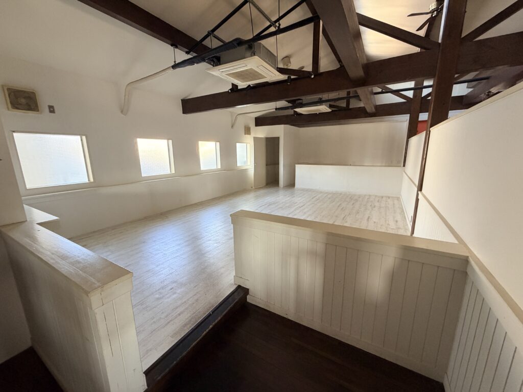 貸店舗の広々とした室内フロアの内観写真。天井は高く、温かみのある太い木製の梁(はり)がむき出しになった開放的なデザイン。白を基調とした壁には小窓が並び、明るい木目調のフローリング床に自然光が差し込んでいる。手前と奥で緩やかに空間が区切られており、カフェやスタジオ、お洒落なオフィスに適した居抜き・スケルトン風テナントの内装。