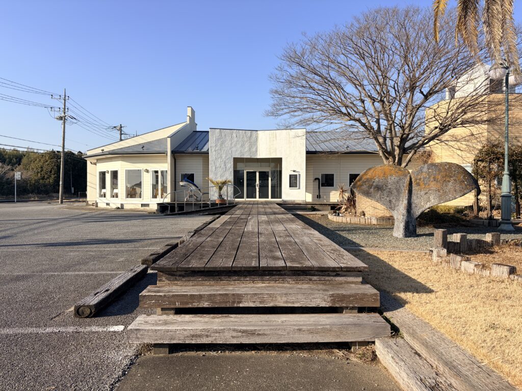 晴天の下、店舗正面へと真っ直ぐに伸びる木製のウッドデッキ通路(アプローチ)を正面から捉えた、平屋建て貸店舗の外観写真。右手前にはクジラの尾の形をした大きな石のオブジェやヤシの木があり、左手にはアスファルトの駐車場が広がっている。海辺のリゾートを思わせる、個性的で開放的なデザインのテナント物件。