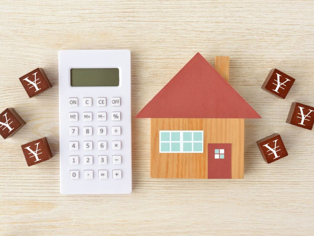 木目調の背景に、赤い屋根の家の模型と白い電卓、そして日本円の記号（¥）が書かれた木製ブロックが並ぶ、住宅ローンの資金計画や計算をイメージした写真