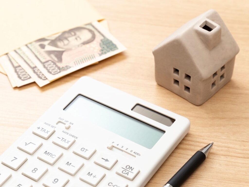 木目調のテーブルの上に、白い電卓、家を模したセラミック製の模型、そして封筒から少し出た日本円の10,000円札(諭吉像が描かれた紙幣)が配置されている。手前には黒いペンも置かれている。日本の住宅購入資金や住宅ローンの計算を連想させるシーン。